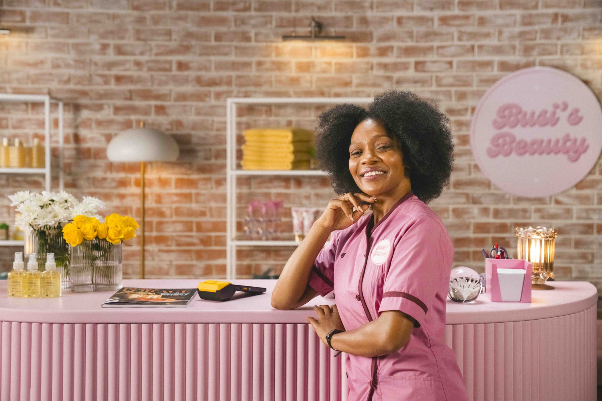 Smiling salon owner in a pink uniform leaning on a pastel‑pink reception desk topped with flowers, beauty products, a magazine and a card reader, with a ‘Busi’s Beauty’ sign against a brick‑wall backdrop.
