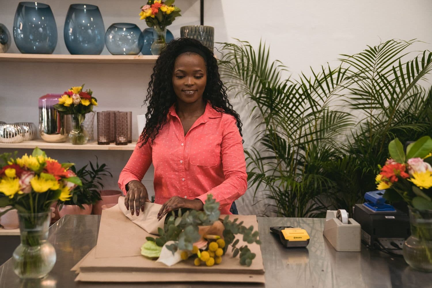 Woman in a coral shirt wrapping a mixed bouquet of flowers and greenery in brown craft paper at a metal counter, with glass vases of flowers on shelves and potted plants in the background.