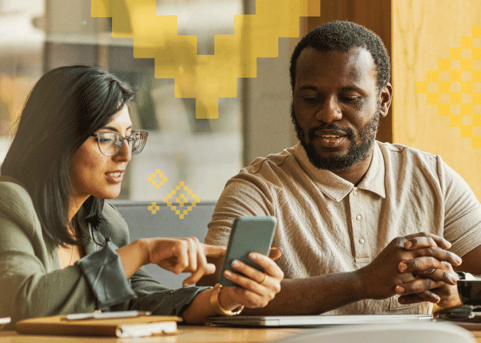 Two colleagues sitting at a table looking at a smartphone, discussing work with notebooks and a laptop in front of them.