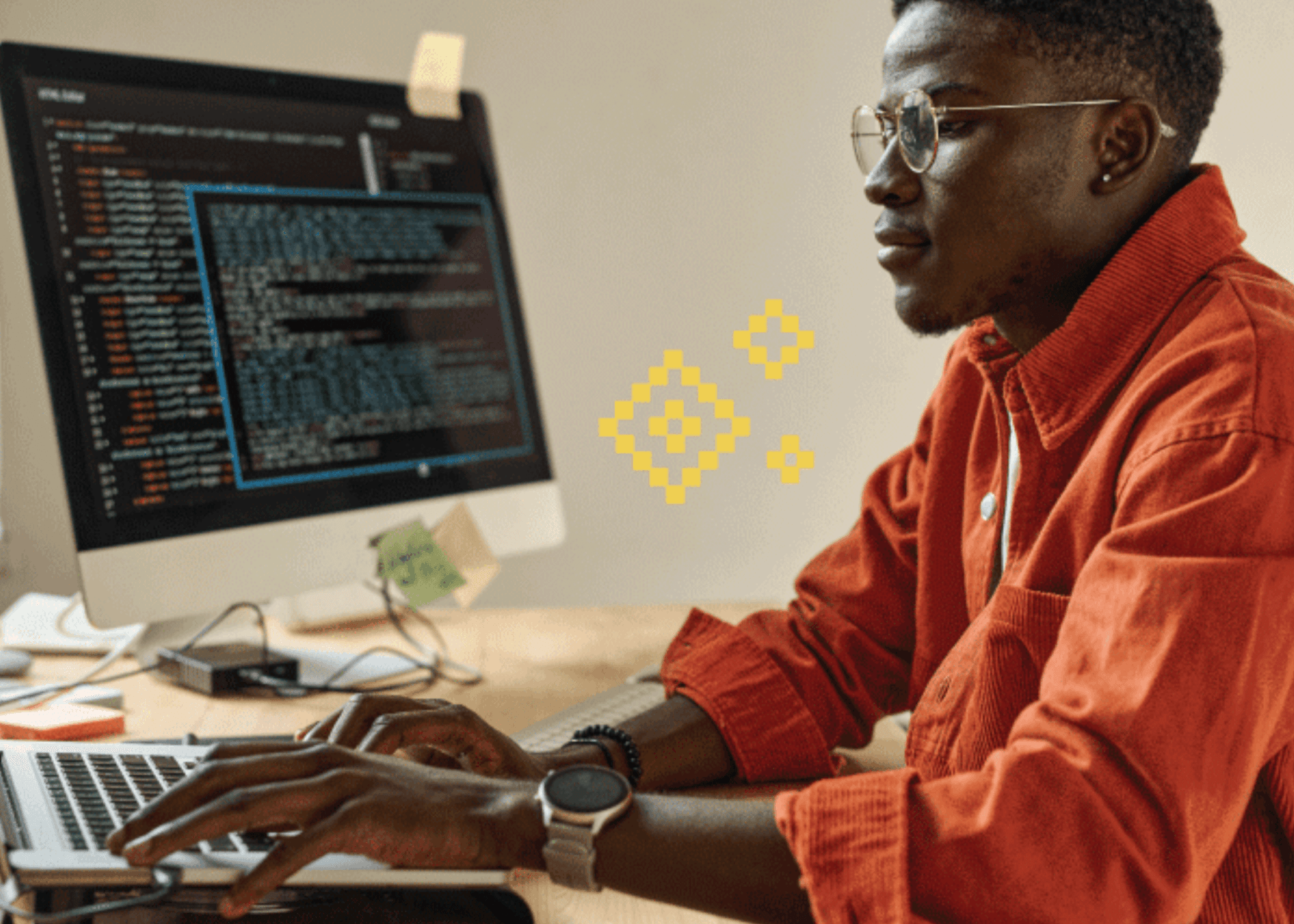 Man wearing glasses and an orange shirt working on a computer with code displayed on the screen.
