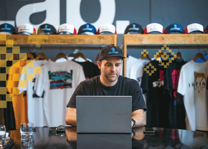 Man wearing a cap working on a laptop in a clothing store with t-shirts and caps displayed behind him.
