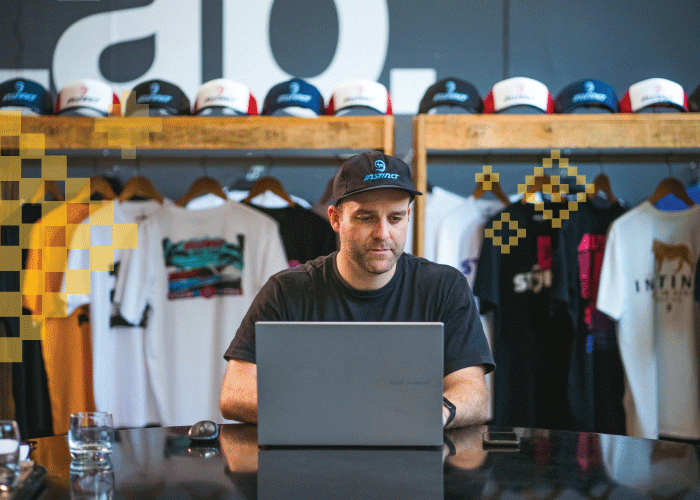 Man wearing a cap working on a laptop in a clothing store with t-shirts and caps displayed behind him.
